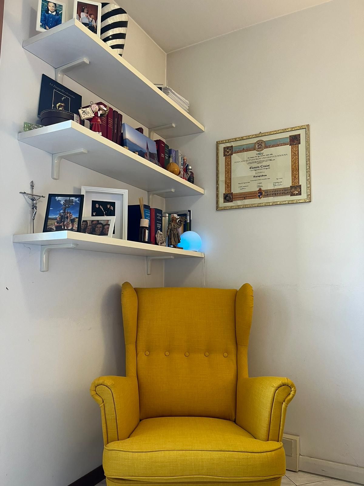 Poltrona gialla in un angolo con scaffali sospesi su cui sono esposti libri e opere d'arte, accanto a un documento incorniciato.
