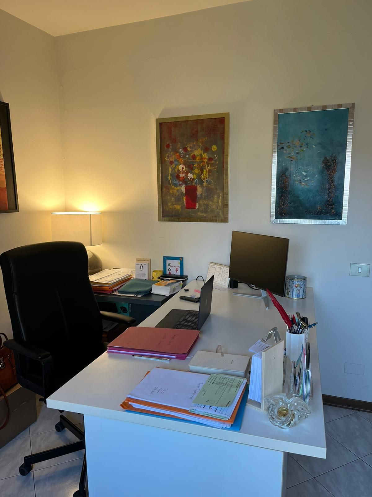 Scrivania da ufficio con computer portatile, documenti e quadri alla parete.