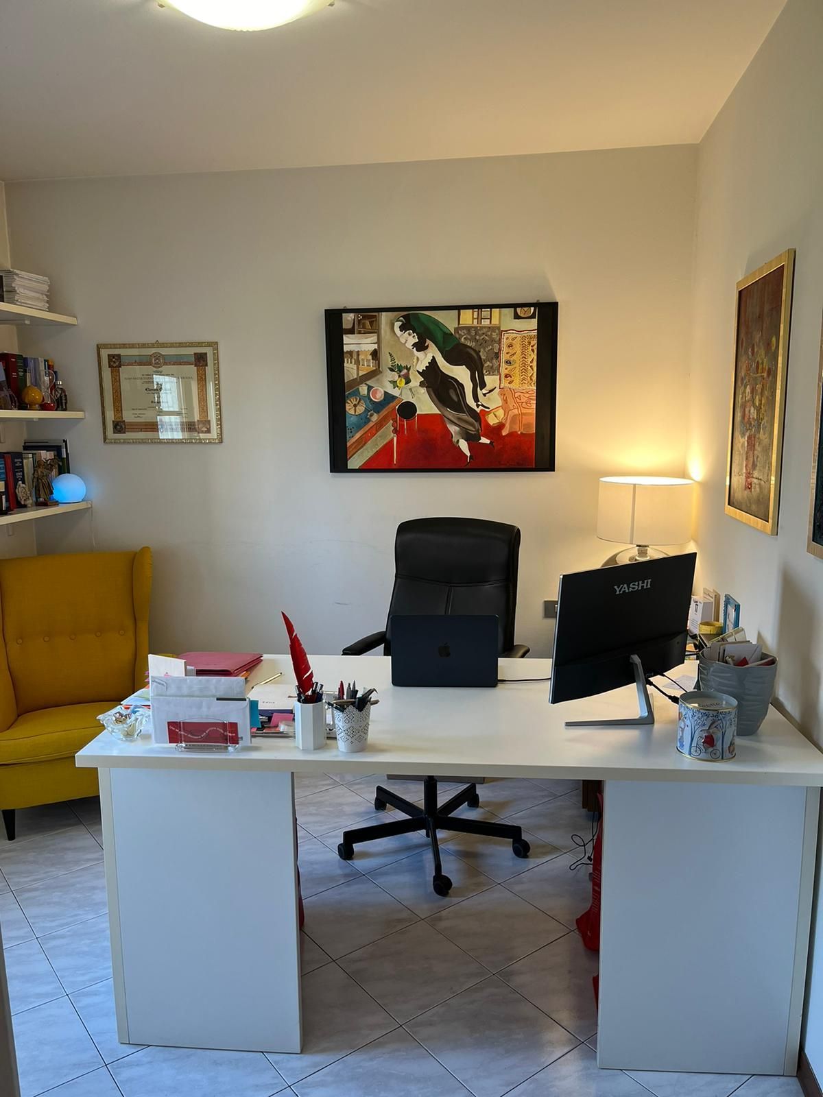 Ufficio bianco con scrivania, sedia, quadri, poltrona gialla e scaffali con libri.