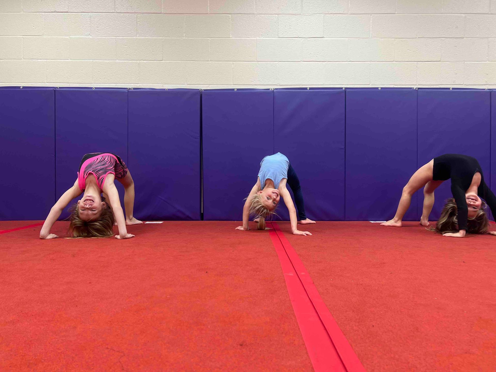 Three young girls are doing a handstand on a red mat in a gym.