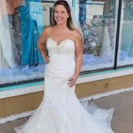 A smiling person in a strapless, floor-length white wedding dress with a lace bodice, posing in front of a shop window.