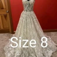 A white, lace-detailed A-line wedding dress on a mannequin in front of a red wall, with the text 