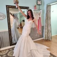 A bride in a white mermaid-style wedding dress holds a bouquet overhead, smiling in front of a mirror in a bridal shop.