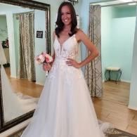 A person wearing a white, A-line wedding dress with floral lace detailing, holding a small bouquet of pink flowers.