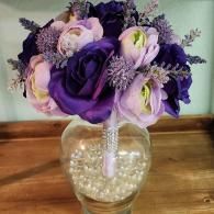 A bouquet of purple and light lavender roses with filler flowers, set in a clear vase filled with white pearl beads.