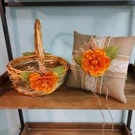A woven wicker basket and a burlap ring pillow, both decorated with white lace and bright orange flowers, on a shelf.