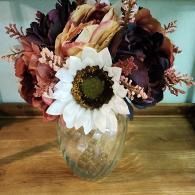 A bouquet of faux burgundy, peach, and white flowers in a clear, fluted glass vase sitting on a wooden surface.