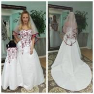 A person and a child wearing matching white, floral-embroidered gowns and veils with dark red trim in a dressing room.