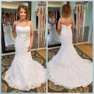 A person wearing a white, strapless, lace mermaid-style wedding dress, shown from the front and back in a bridal shop.