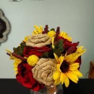 A bouquet of red silk flowers, bright yellow sunflowers, and tan burlap roses arranged in a rustic, autumnal style.