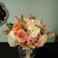 A lush, artificial wedding bouquet featuring peach, pink, and cream roses with greenery in a crystal-embellished vase.