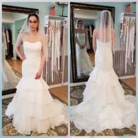 A woman wearing a white, tiered, strapless mermaid-style wedding gown and veil, shown from the front and back in a mirror.