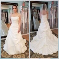 A person stands in a bridal shop, wearing a strapless white wedding gown with ruched detailing and a veil.