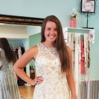 A smiling person wearing a sleeveless, heavily beaded white formal gown in a clothing boutique.