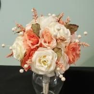A bridal bouquet of peach and cream roses accented with white pearl beads and dried foliage in a glass vase.