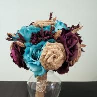 A wedding bouquet of teal and deep plum silk roses mixed with burlap roses in a glass vase against a white wall.