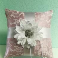 A square, dusty pink ring bearer pillow featuring white lace overlay, a central white fabric flower, and a silver ribbon.