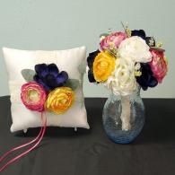 A wedding ring pillow with silk flowers sits next to a matching bouquet in a glass vase on a dark surface.