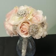 A bridal bouquet of light pink and cream roses adorned with rhinestone brooches, held in a clear glass vase.