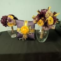 A purple ring pillow featuring a yellow flower, flanked by two vases of yellow and dark purple artificial floral bouquets.