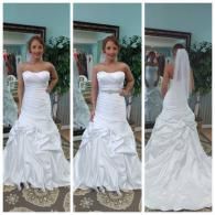 A woman in a bridal shop wearing a white, strapless, mermaid-style wedding gown with ruffles and a veil in the back view.