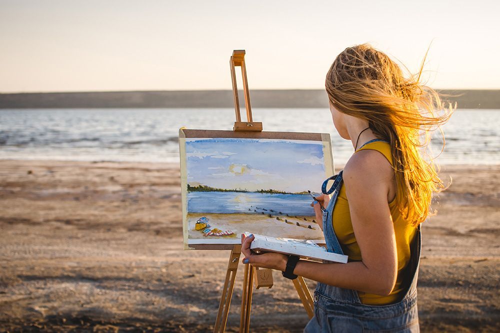 Woman painting a watercolor landscape at the beach; sunset light, easel, brush, palette.