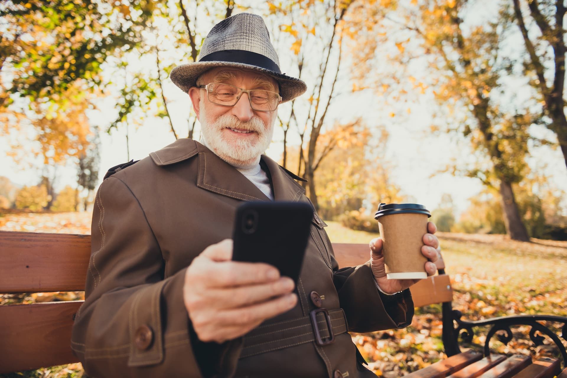 Man in a hat and coat looks at phone while holding a coffee cup in a park with autumn foliage.