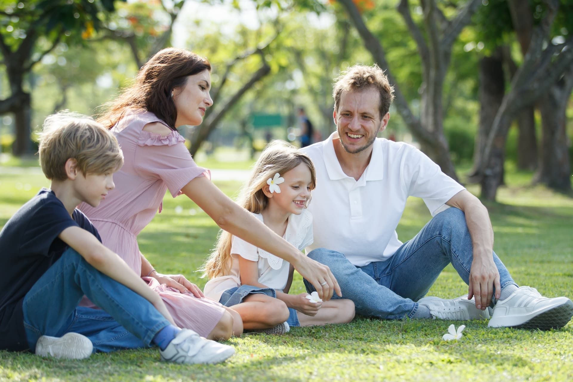Family sits on grass in park, interacting. Woman in pink dress, man in white shirt, two children. Sunny day.