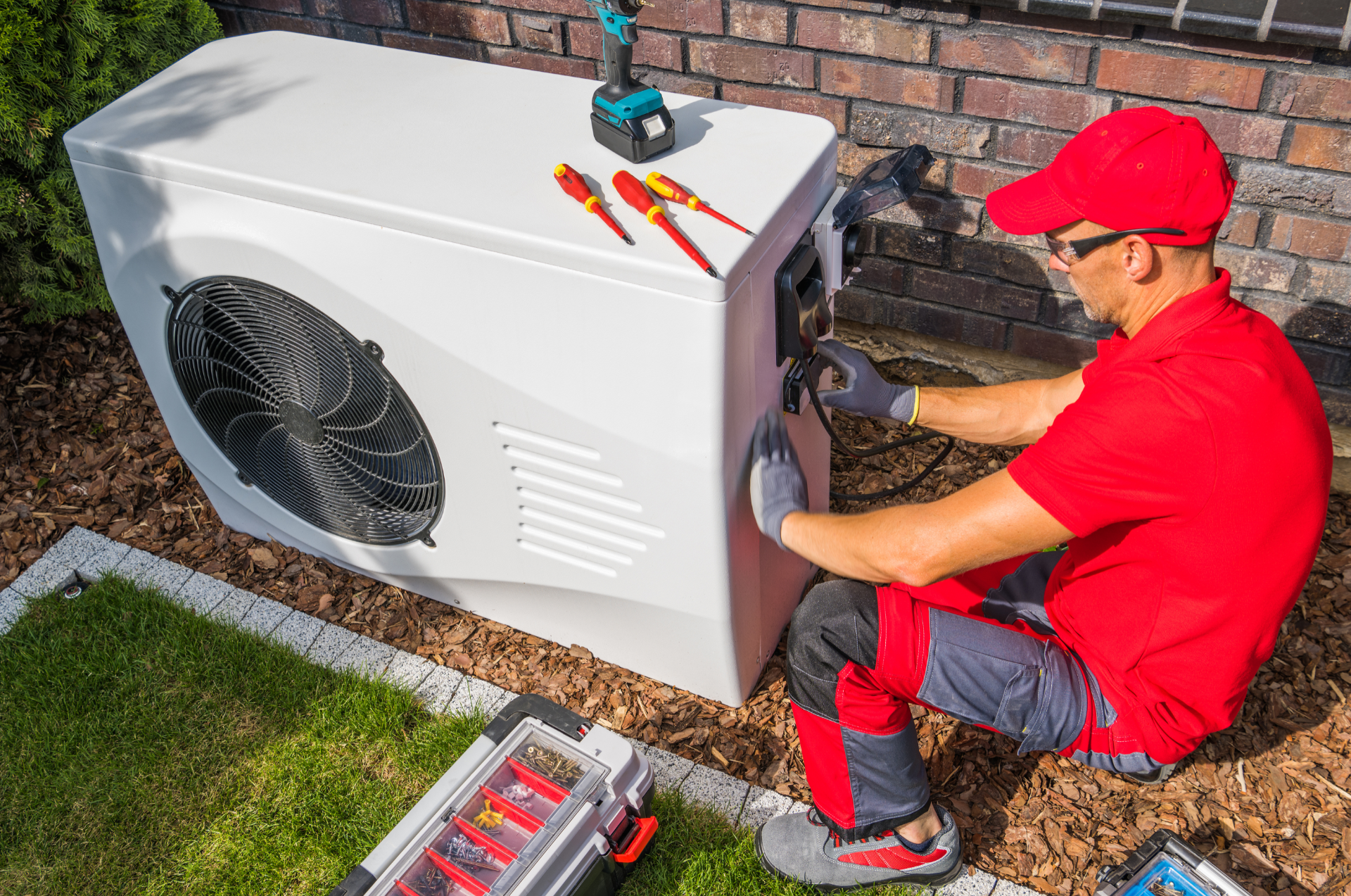 Technician in red shirt working on a white heat pump outside a brick building.