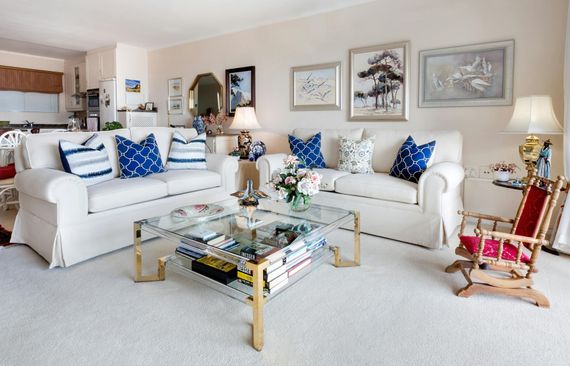 Living room with white couches, blue pillows, glass coffee table, art, and rocking chair.