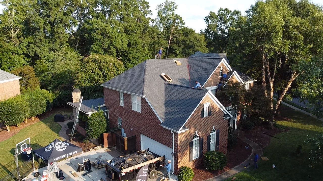 Two-story brick house with a dark roof surrounded by green trees.