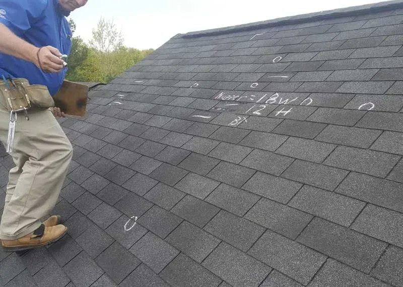 Person inspecting a dark gray shingle roof, with white chalk marks.