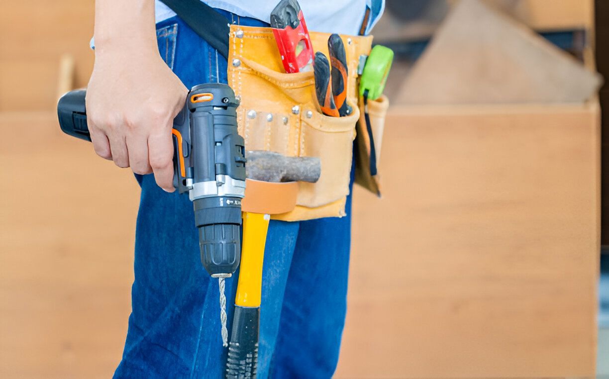 Person in jeans, holding a drill, tool belt with hammer, gloves, and measuring tape.