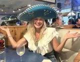A smiling person wearing a large teal sombrero and a ruffled white top, sitting at a restaurant table with hands raised.