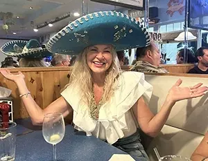 A smiling person wearing a large teal sombrero and a ruffled white top, sitting at a restaurant table with hands raised.