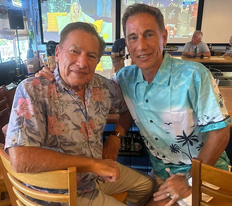 Two men in short-sleeved, patterned shirts smile while sitting side-by-side at a wooden table in a brightly lit venue.