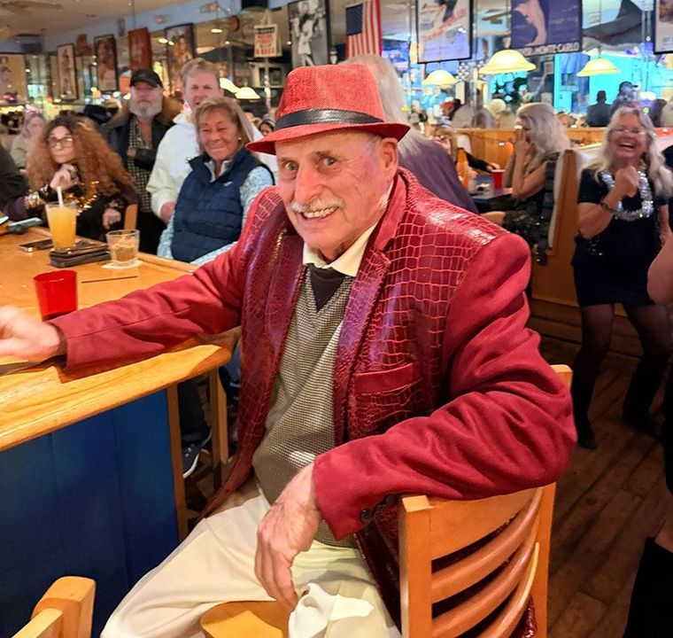 A man in a textured red blazer and matching fedora sits smiling at a bar, with people blurred in the background.