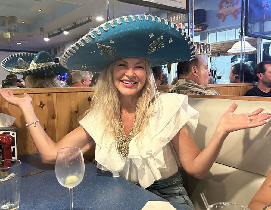 A smiling person wearing a large, teal, decorative sombrero at a restaurant table with their arms outstretched.