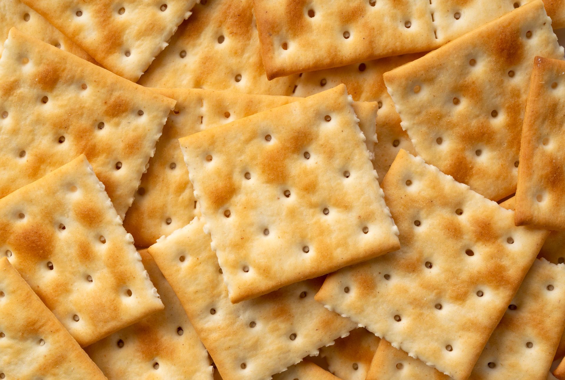 Mucchio di cracker quadrati, dorati, con la superficie forata, sparsi uno vicino all'altro.