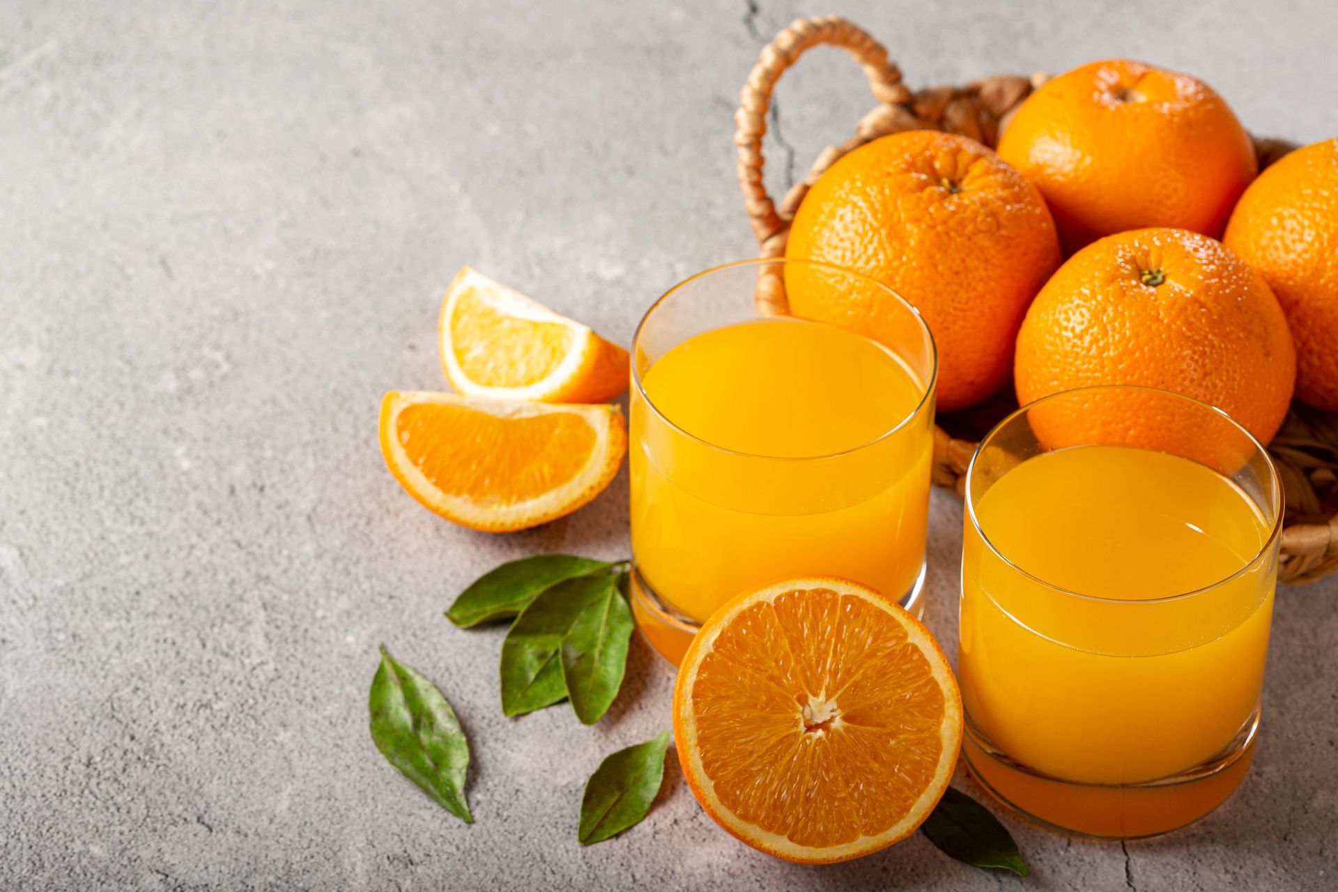 Due bicchieri di succo d'arancia con arance in un cestino su una superficie grigia con fette e foglie d'arancia.