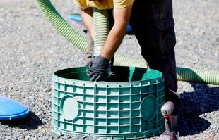 A worker in dark pants and gloves uses a large green hose to service a green plastic septic tank riser on gravel ground.