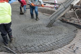 Construction workers use tools to smooth wet concrete pouring from a chute onto a metal grid base.
