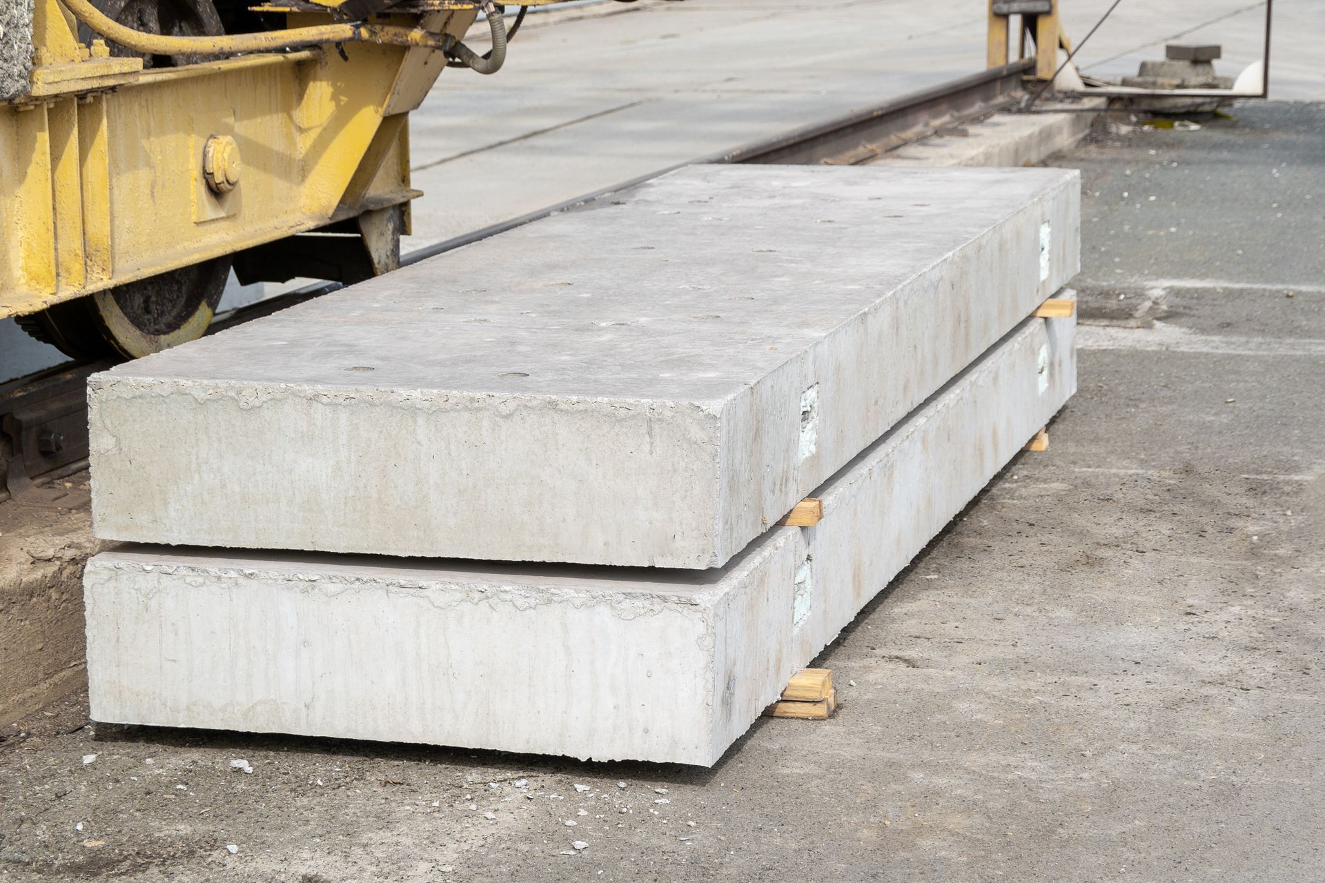 Two large, rectangular concrete slabs stacked on wooden blocks outdoors, next to a piece of yellow construction equipment.