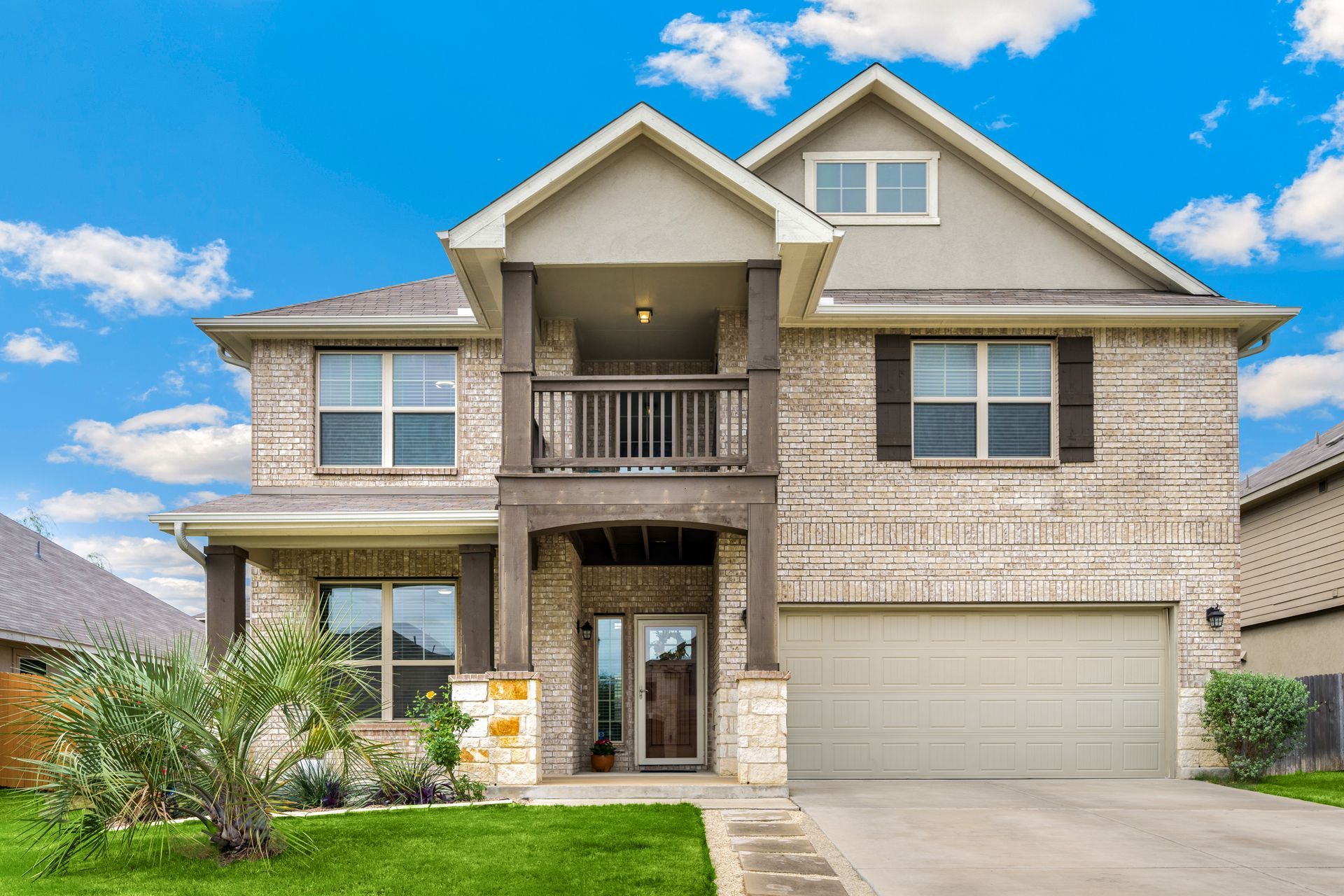 A two-story tan brick house with a front porch, a balcony above the entryway, and a two-car garage under a blue sky.