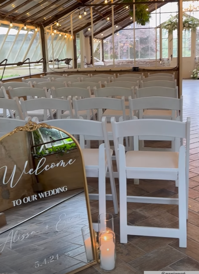 A wedding ceremony is taking place in a greenhouse with white chairs and a welcome sign.