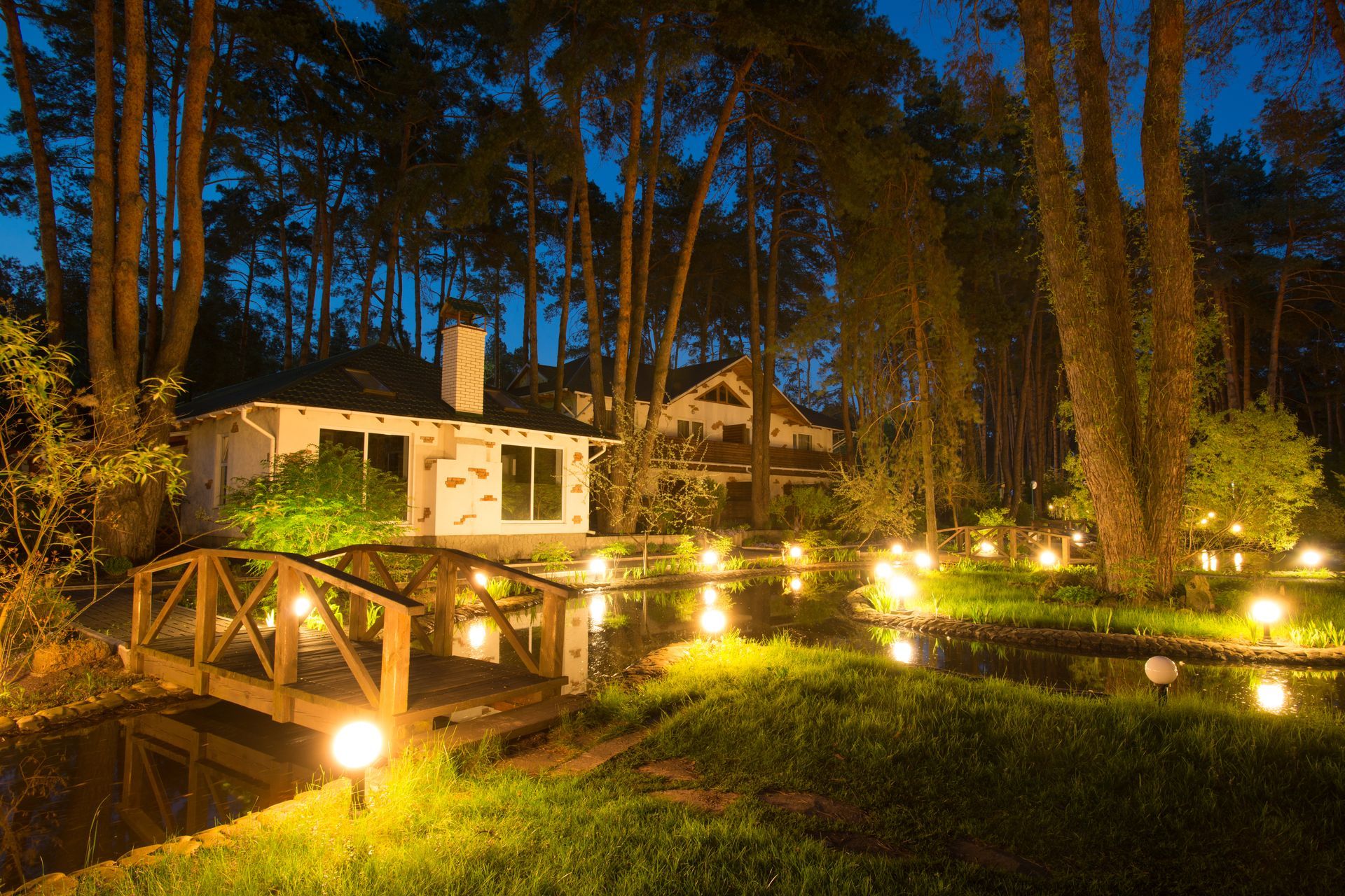 Nighttime garden with a lit wooden bridge, warm pathway lights, and a house among tall trees.