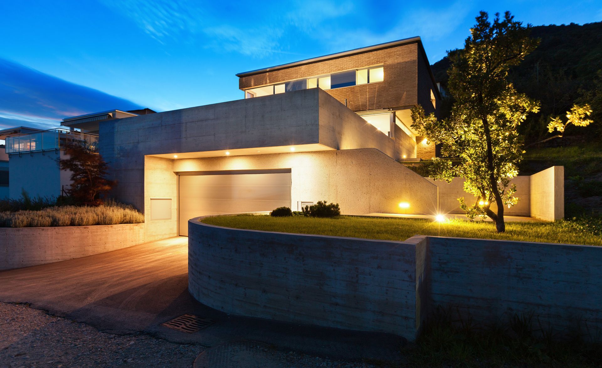 Modern house at dusk with warm exterior lighting, garage, and curved stone wall in front yard