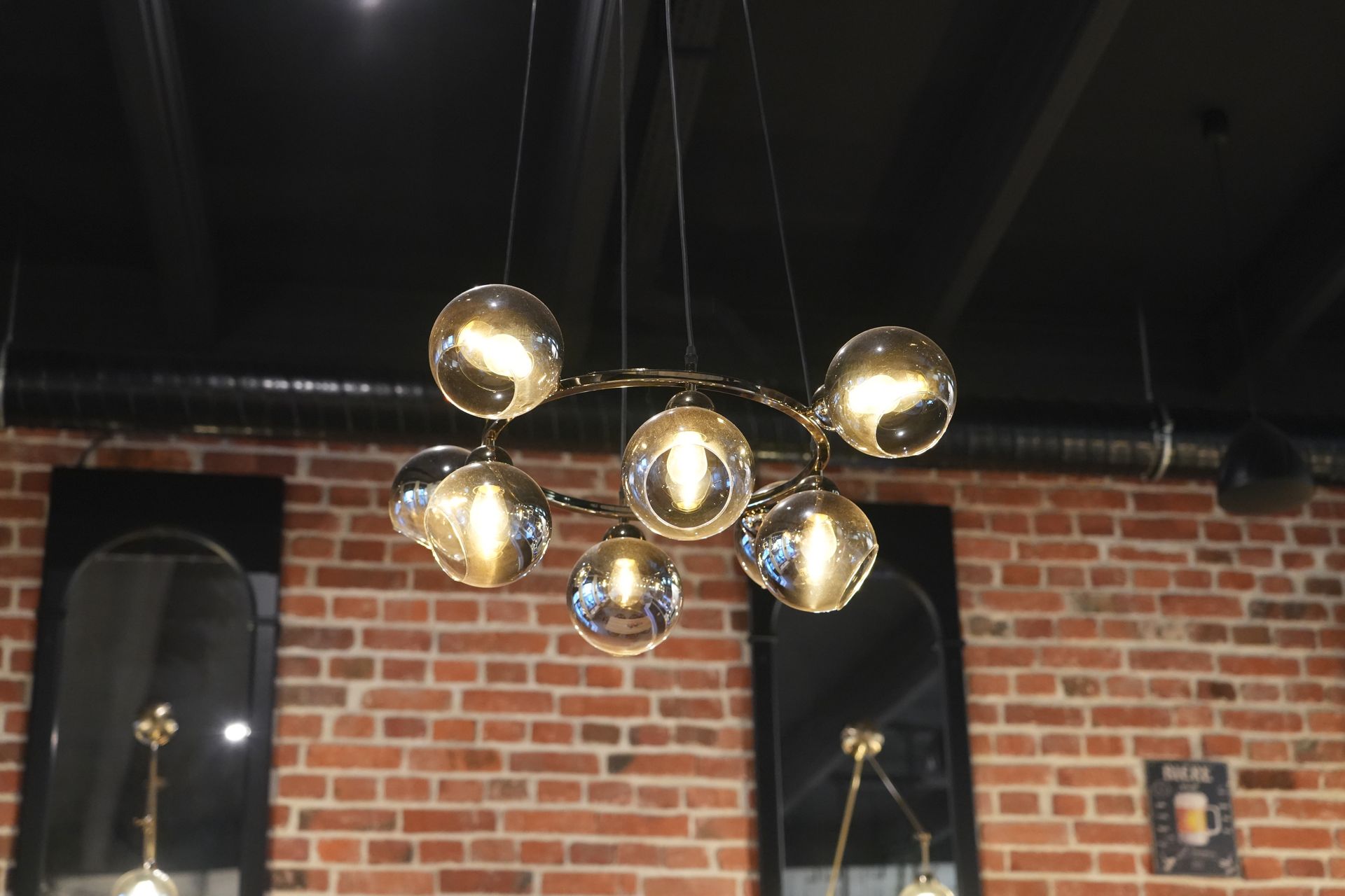 Modern gold globe chandelier hanging from a black ceiling above a brick wall