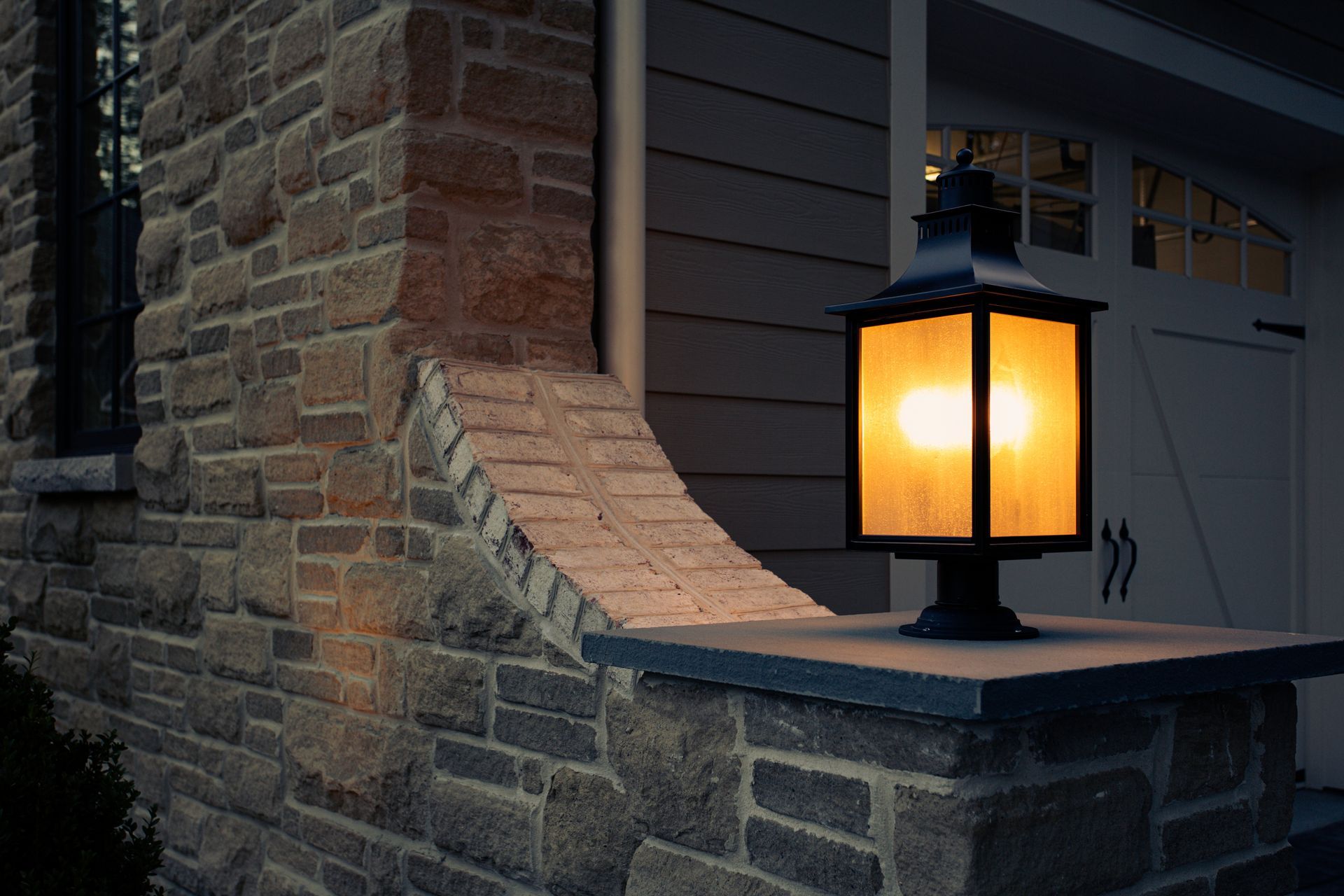 Glowing black lantern on a stone porch beside a brick wall and garage at dusk