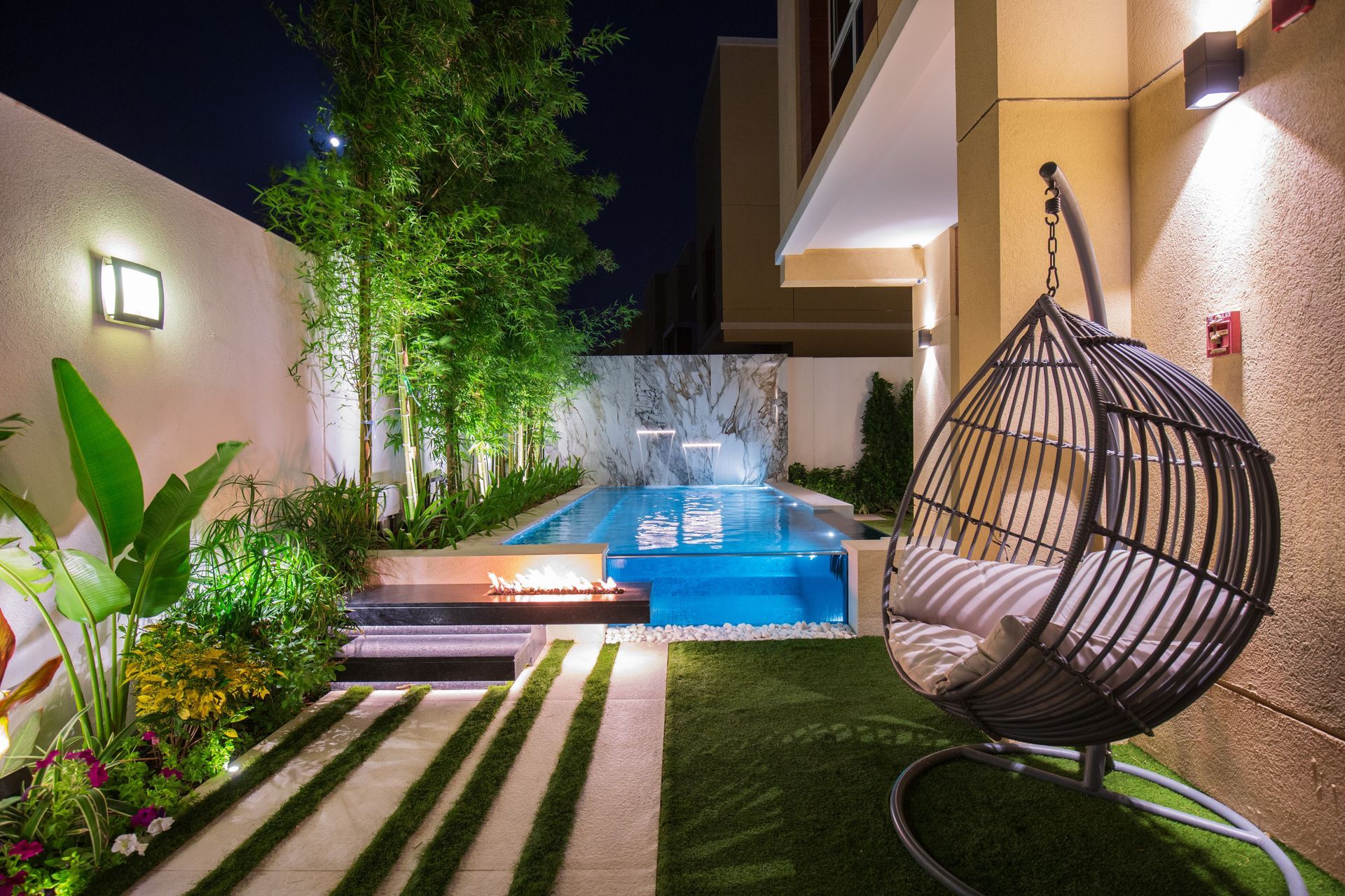 Nighttime backyard with pool, hanging chair, and lush landscaping, illuminated by warm lighting.